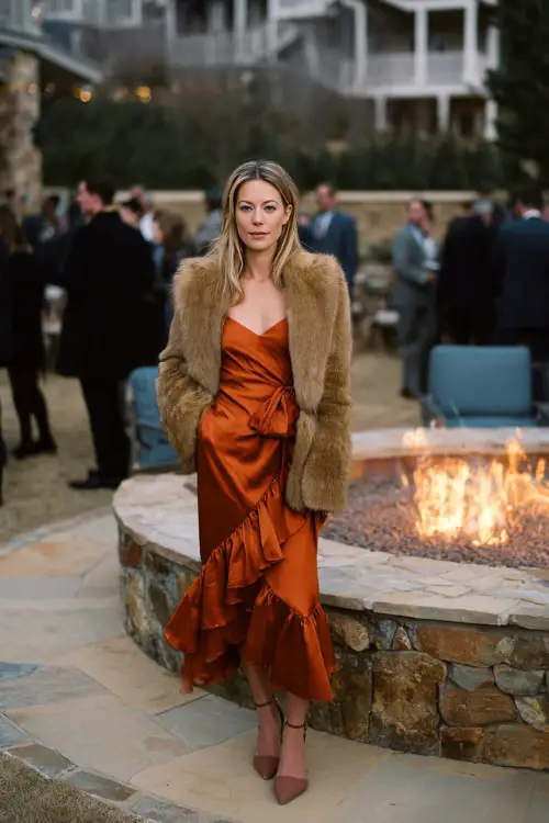 A woman in her 30s wears a rust-colored ruffled midi dress with a tan fur coat