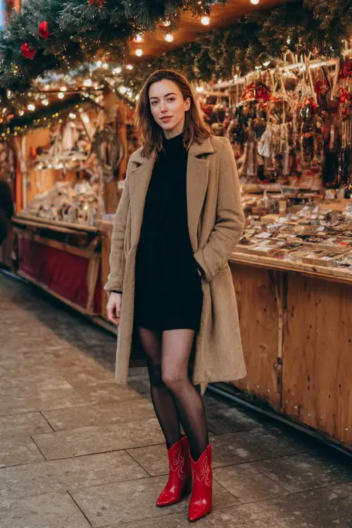 A woman over 30 wears a black mini dress with black tights and short red cowboy boots, layered with a camel coat