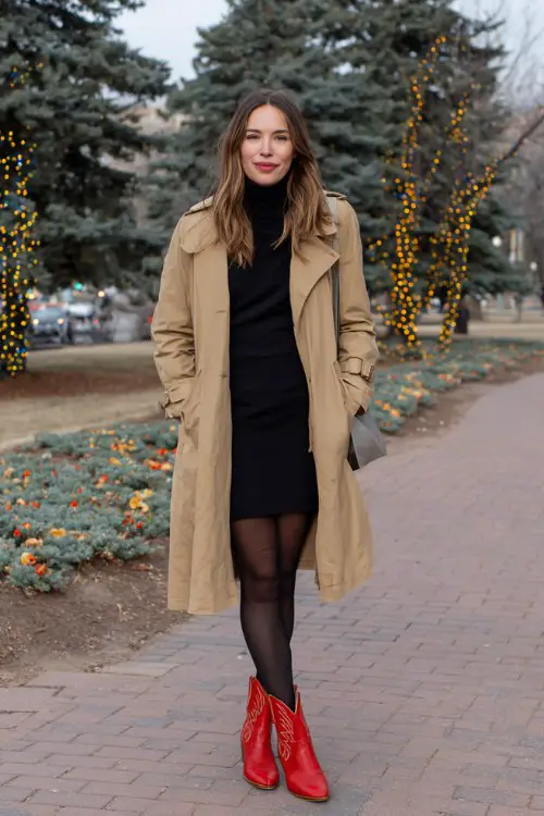 A woman over 30 wears a black turtleneck dress with black tights and red cowboy boots, layered with a camel trench coat