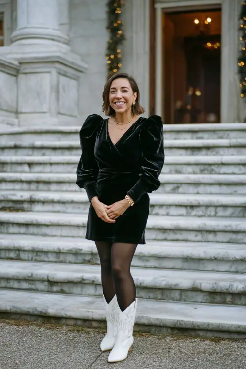 A woman over 30 wears a black velvet dress with balloon sleeves, black tights, and white cowboy boots