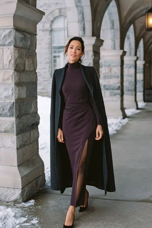 A woman over 30 wears a dark plum high-neck gown with long fitted sleeves, side slit, and sheer black tights, layered with a tailored wool cape coat and black pumps