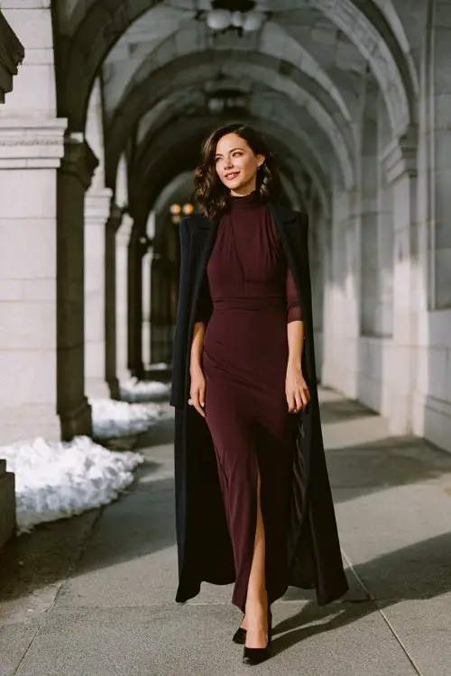A woman over 30 wears a dark plum high-neck gown with long fitted sleeves, side slit, and sheer black tights, layered with a tailored wool cape coat and black pumps