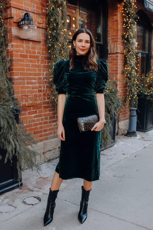 A woman over 30 wears a deep emerald velvet mock-neck midi dress with puffed sleeves and pointed leather boots