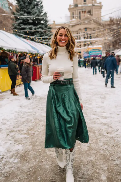 A woman over 30 wears a green satin midi skirt with a cream turtleneck and white cowboy boots