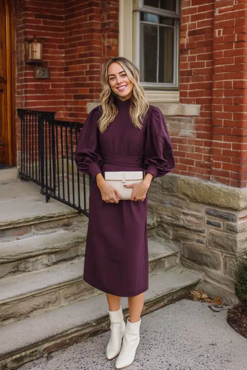 A woman over 30 wears a plum midi dress with balloon sleeves and a mock neck, and cream sock boots, holding a small clutch