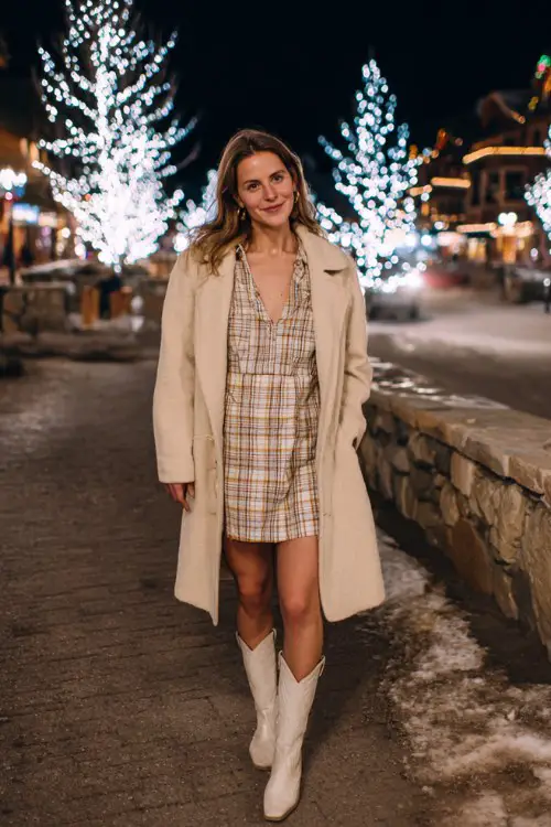 A woman over 40 wears a plaid mini dress layered under a long beige wool coat with white cowboy boots
