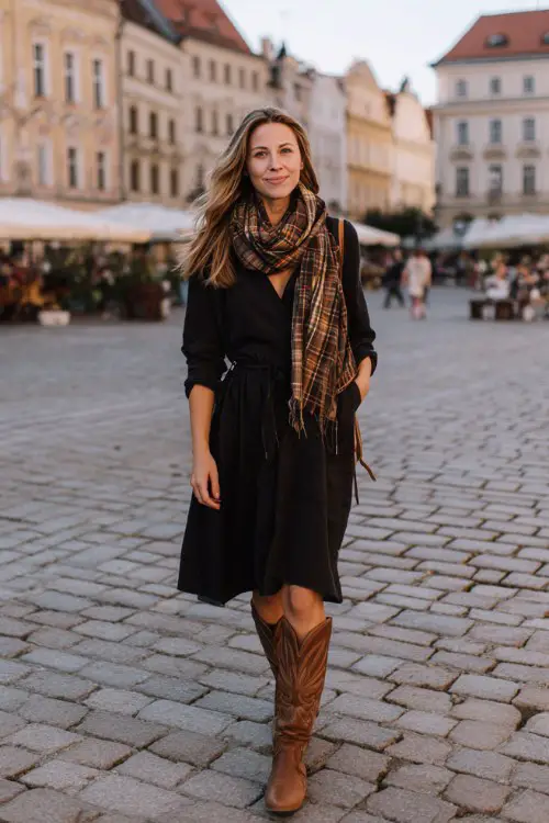 A woman over 40 wears brown tall cowboy boots with a black knit dress and a plaid scarf