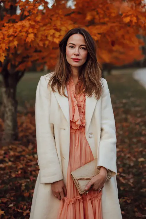 A woman wears a blush chiffon dress layered with a cream wool coat