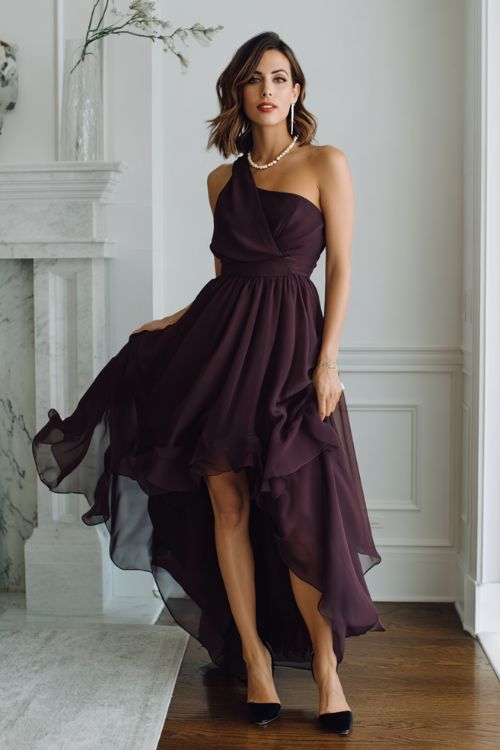 A woman wears a dark plum chiffon gown with layered flowing fabric and sleek black stilettos, styled with pearl accessories