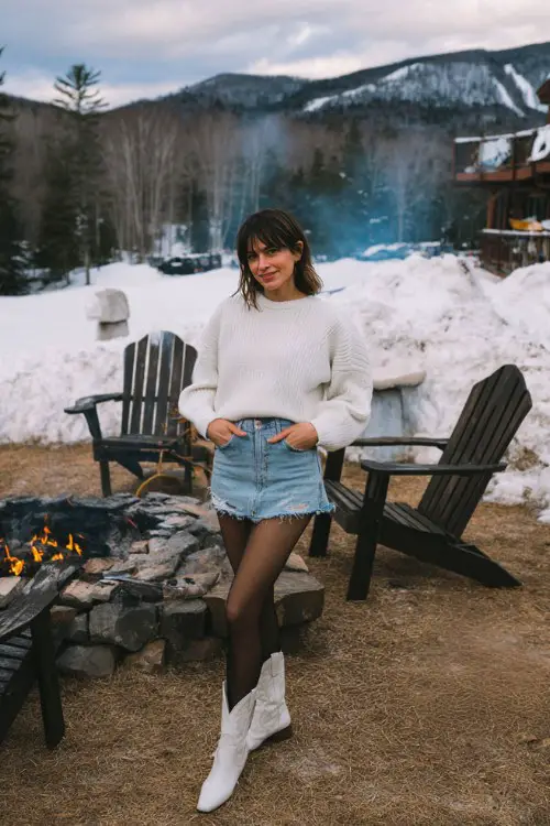 A woman wears a denim mini skirt with black tights, a white sweater, and short white cowboy boots