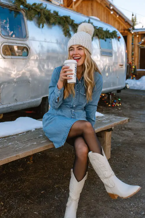 A woman wears a fitted denim shirt dress with black tights, white cowboy boots, and a cozy knit beanie