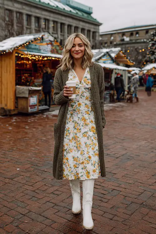 A woman wears a floral midi dress layered with a longline cardigan and tall white cowboy boots
