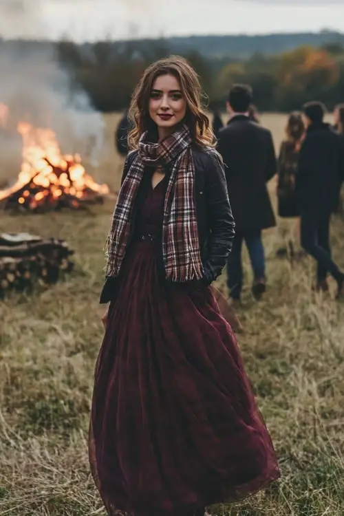 A woman wears a maroon dress with a plaid scarf and jacket, attending a rustic October wedding with a bonfire in the background