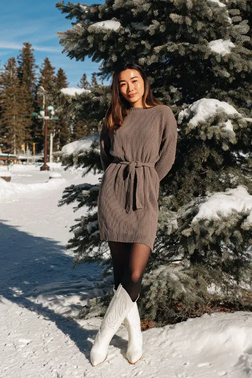A woman wears a mocha ribbed sweater dress cinched at the waist, paired with black tights and white cowboy boots
