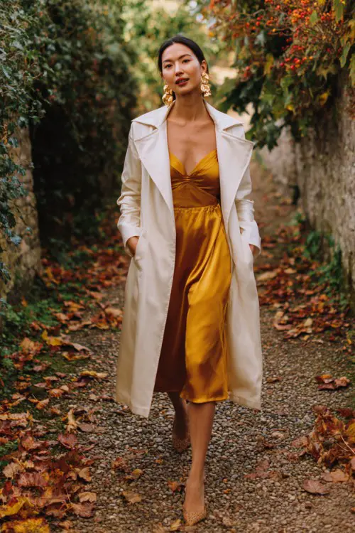 A woman wears a mustard satin dress with a long ivory coat and gold earrings