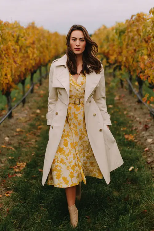 A woman wears a mustard yellow floral tea-length dress with a belted cream trench coat 