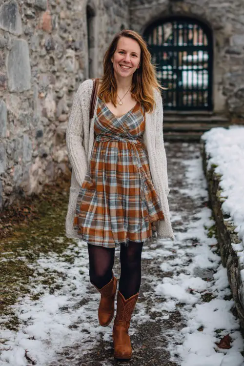 A woman wears a plaid winter dress in warm tones with black tights and brown cowboy boots, layered with a soft cardigan