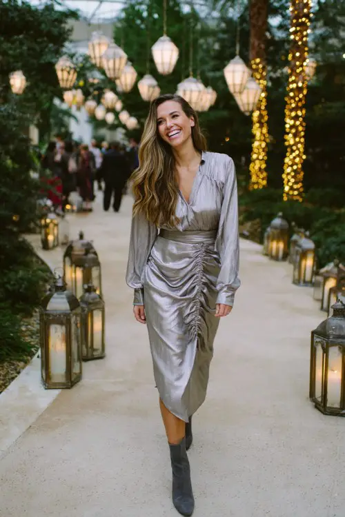 A woman wears a silver metallic long-sleeve midi dress with subtle shimmer and sculptural draping, paired with dark gray suede booties