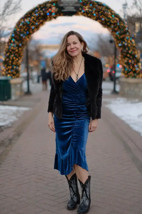 A curvy woman over 40 wears a royal blue velvet midi dress with a black faux fur wrap and black cowboy boots with silver detailing