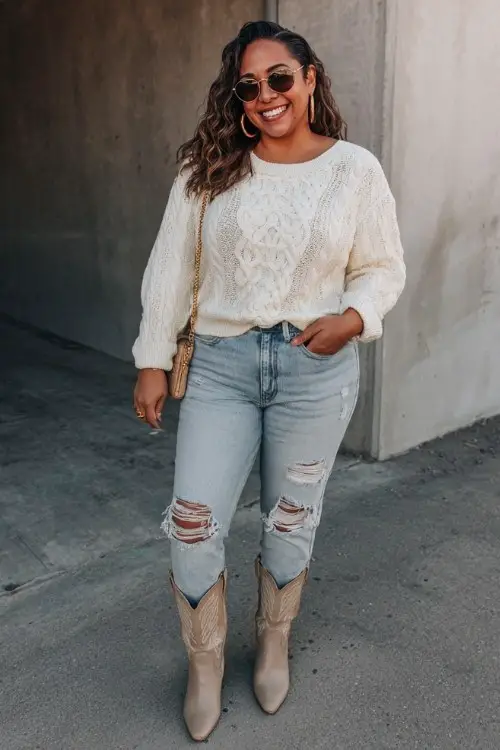 A curvy woman rocks a cream cable-knit sweater, ripped light-wash jeans, and beige cowboy boots