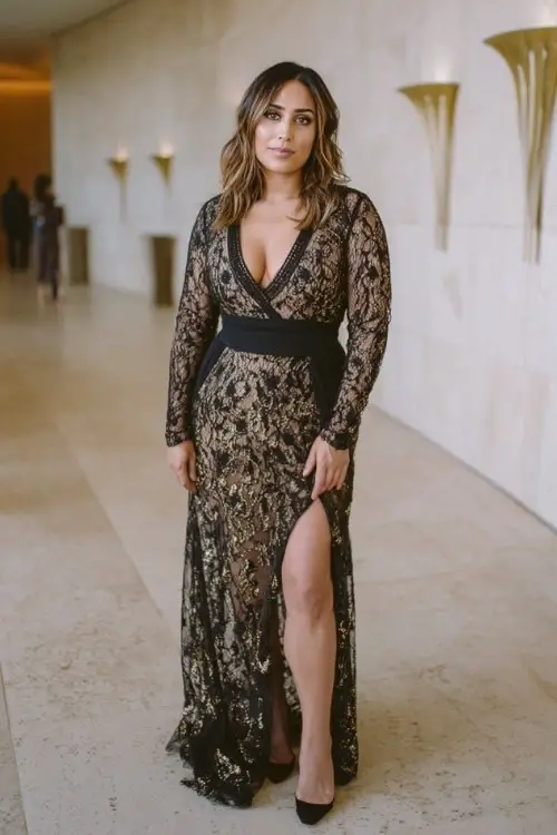 A curvy woman wears a black base gown with gold lace overlay and long illusion sleeves, styled with black suede heels and elegant studs