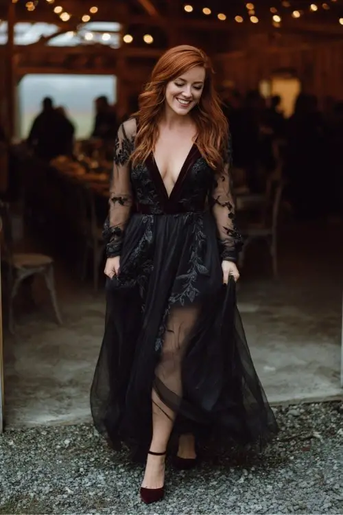 A curvy woman wears a black gown with moody floral embroidery, sheer long sleeves, and a flowing tulle skirt, styled with burgundy heels and a velvet wrap