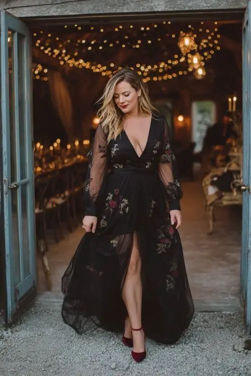 A curvy woman wears a black gown with moody floral embroidery, sheer long sleeves, and a flowing tulle skirt, styled with burgundy heels and a velvet wrap