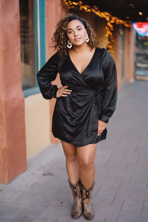 A curvy woman wears a black satin wrap dress with a subtle sheen, styled with two-tone cowboy boots and statement earrings