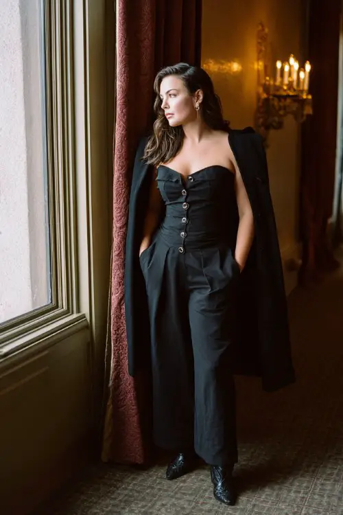 A curvy woman wears a formal black jumpsuit with a cinched waist and flared legs tucked into heeled black cowboy boots