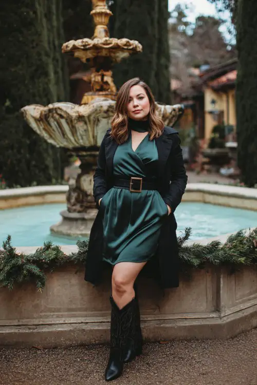 A curvy woman wears a high-neck satin wrap dress in deep forest green, layered with a black belted trench coat and tall black cowboy boots