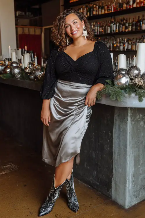 A curvy woman wears a sparkle-accented black top tucked into a satin midi skirt, styled with two-tone cowboy boots and bold earrings