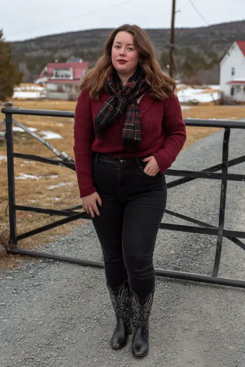 A curvy woman wears high-rise black jeans with embroidered black cowboy boots, a burgundy knit sweater, and a plaid scarf