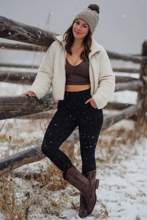 A curvy woman wears thick black leggings with chocolate brown cowboy boots, a cropped ivory sherpa jacket, and a knit hat