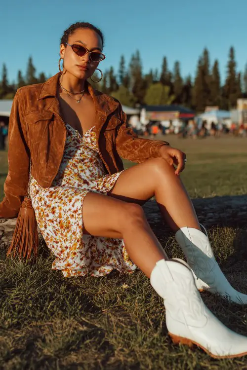 A curvy woman wears white cowboy boots with a floral midi dress layered with a cropped suede jacket, accessorized with hoop earrings and a fringe bag