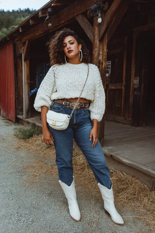 A curvy woman wears white cowboy boots with dark-wash bootcut jeans, a cropped knit sweater and wide western belt