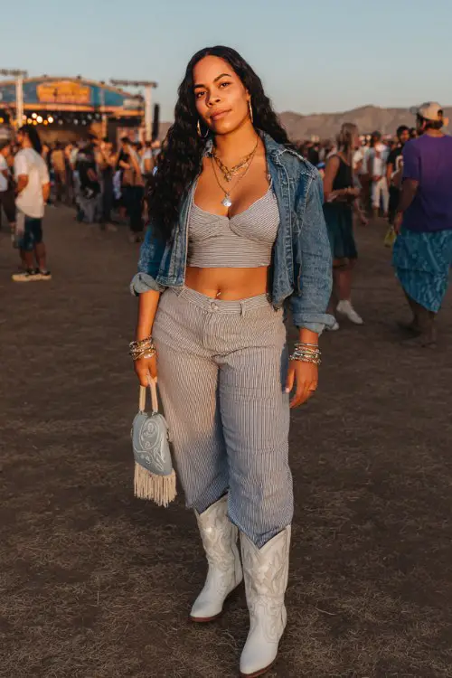 A curvy woman wears white cowboy boots with high-rise wide-leg jeans, a fitted crop tee and cropped denim jacket, accessorized with stacked bracelets and a fringe bag
