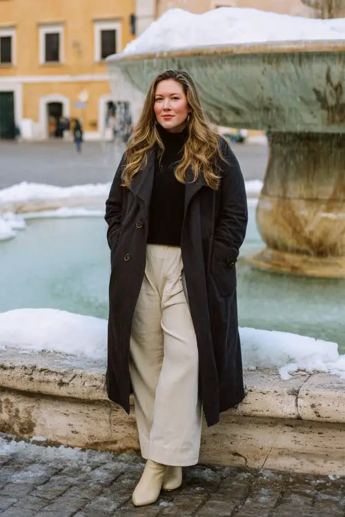 A plus-size woman wears a flowing black trench coat layered over a cream knit turtleneck and wide-leg wool trousers, with short beige cowboy boots