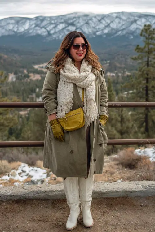 A plus-size woman wears a soft olive green trench coat with a chunky knit scarf, leather gloves, and tall white cowboy boots