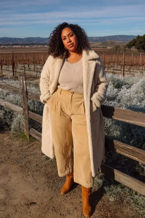 A plus-size woman wears beige wide-leg trousers tucked into tall tan cowboy boots, a knit top, and a long shearling-lined coat