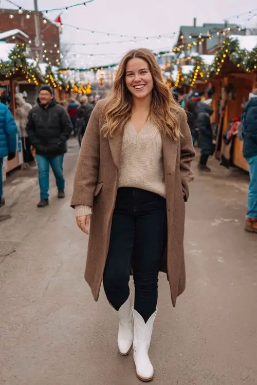 A plus-size woman wears black jeans, a mocha-colored oversized knit top, a structured wool coat, and bright white cowboy boots