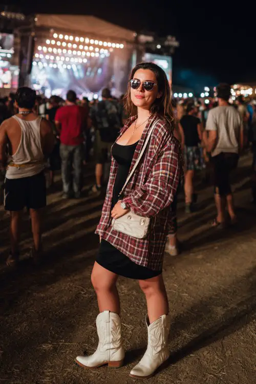 A plus-size woman wears white cowboy boots with a fitted black midi dress and oversized plaid shirt worn open, finished with bold sunglasses and a mini shoulder bag