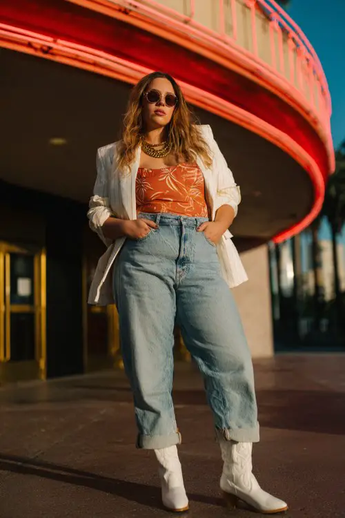 A plus-size woman wears white cowboy boots with light-wash straight jeans, a bold printed bodysuit and oversized blazer, finished with layered gold chains and sunglasses