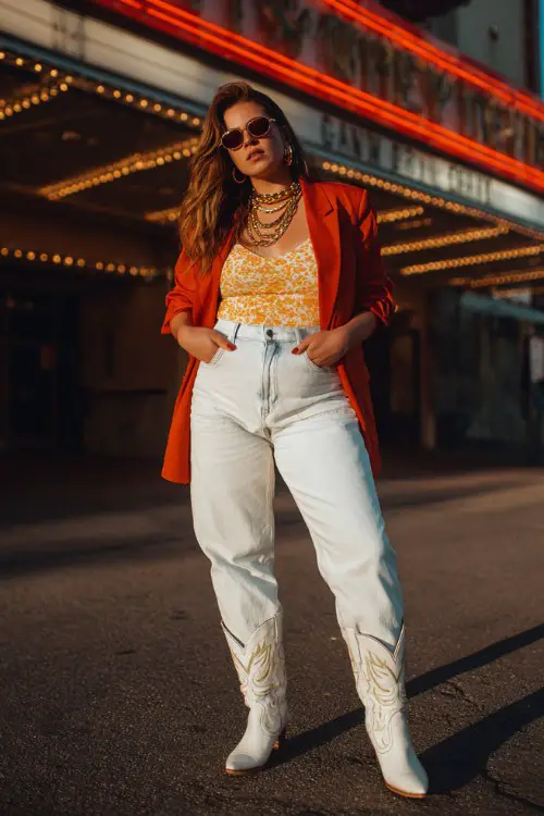 A plus-size woman wears white cowboy boots with light-wash straight jeans, a bold printed bodysuit and oversized blazer, finished with layered gold chains