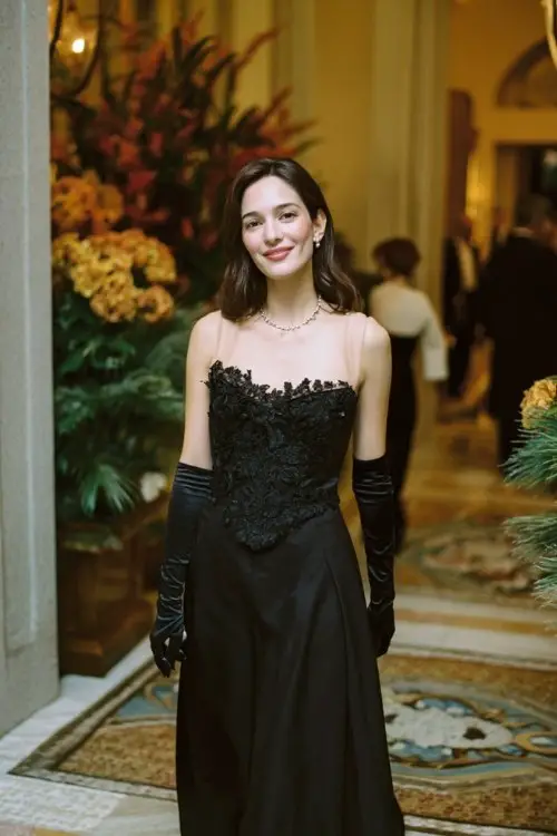 A woman arriving at a winter garden reception in a black A-line gown with a sheer neckline, embroidered bodice, and long velvet gloves