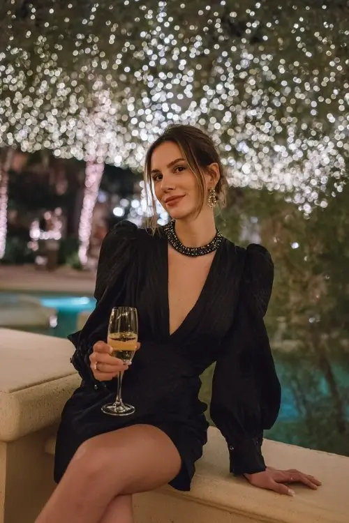 A woman holding a glass of champagne under twinkling string lights in a structured black mini dress with a beaded high neckline and bishop sleeves