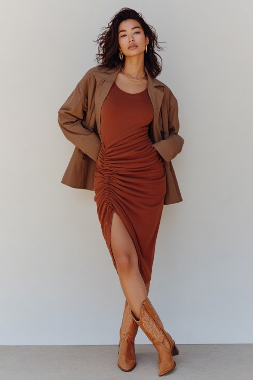 A woman in her 30s wears a cinnamon brown wool midi dress with a subtle slit and ruched waist, styled with caramel cowboy boots