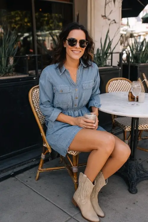 A woman over 30 pairs a chambray romper with beige cowboy boots and oversized sunglasses for a fun outdoor lunch with friends