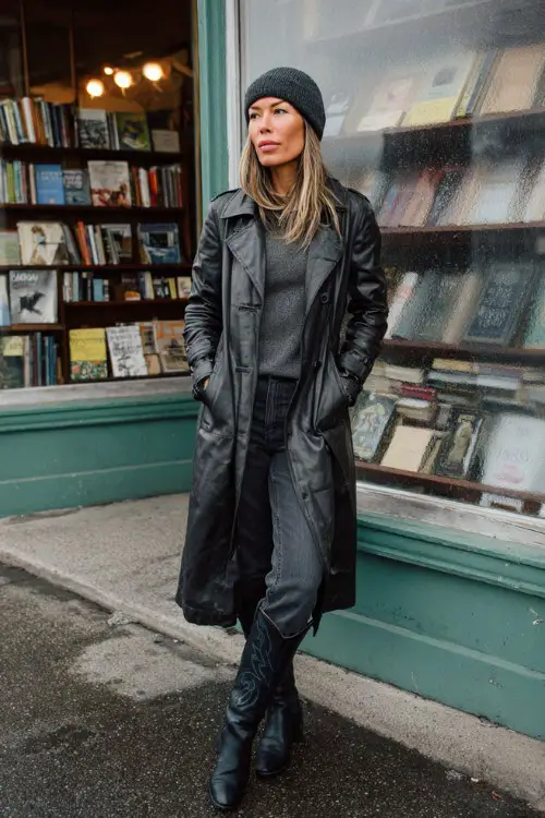 A woman over 30 wears a black leather trench coat layered over dark-wash jeans and a gray knit, paired with tall black cowboy boots and a beanie