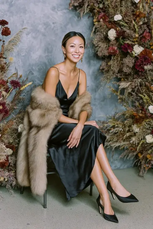 A woman over 30 wears a black satin slip dress layered with a faux-fur stole and pointed-toe heels