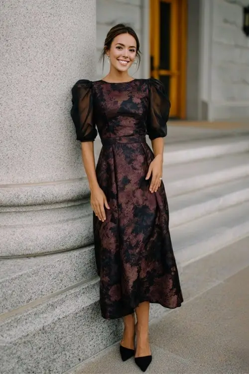 A woman over 30 wears a dark floral brocade gown with sheer puff sleeves and a cinched waist